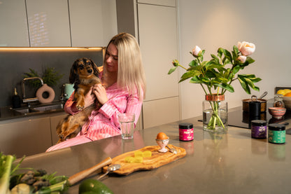 Woman in pink robe holding a dog in a kitchen with flowers and food items on the counter.
