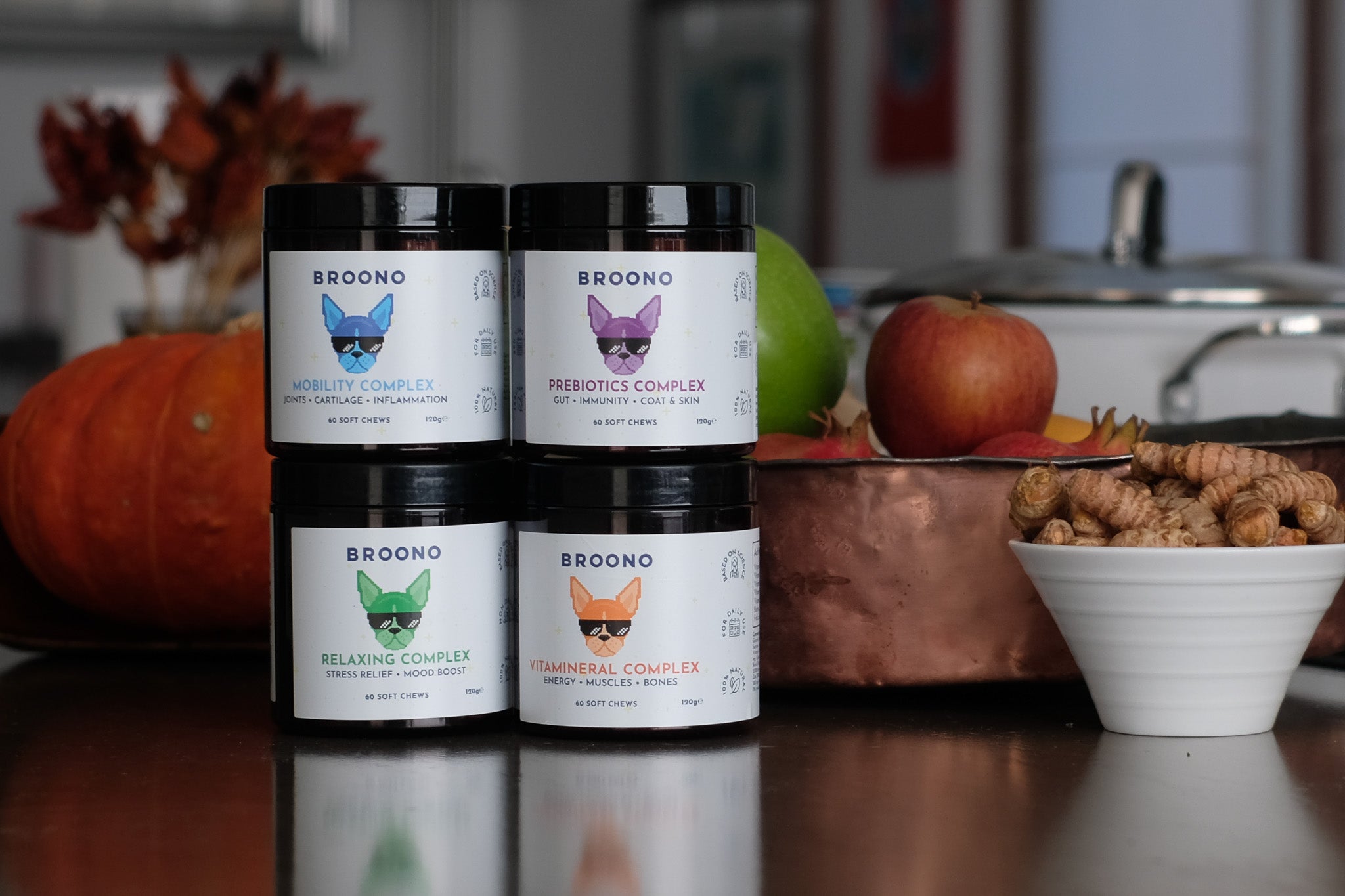 Four Broono pet supplement containers on a kitchen counter with fruits and nuts.